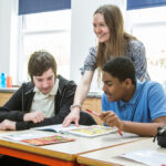 Female teacher from Dawn House smiling and helping two pupils in a classroom.